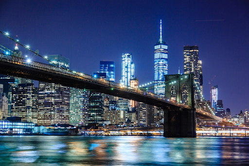 Nighttime skyline of New York City behind the river and iconic Brooklyn bridge.